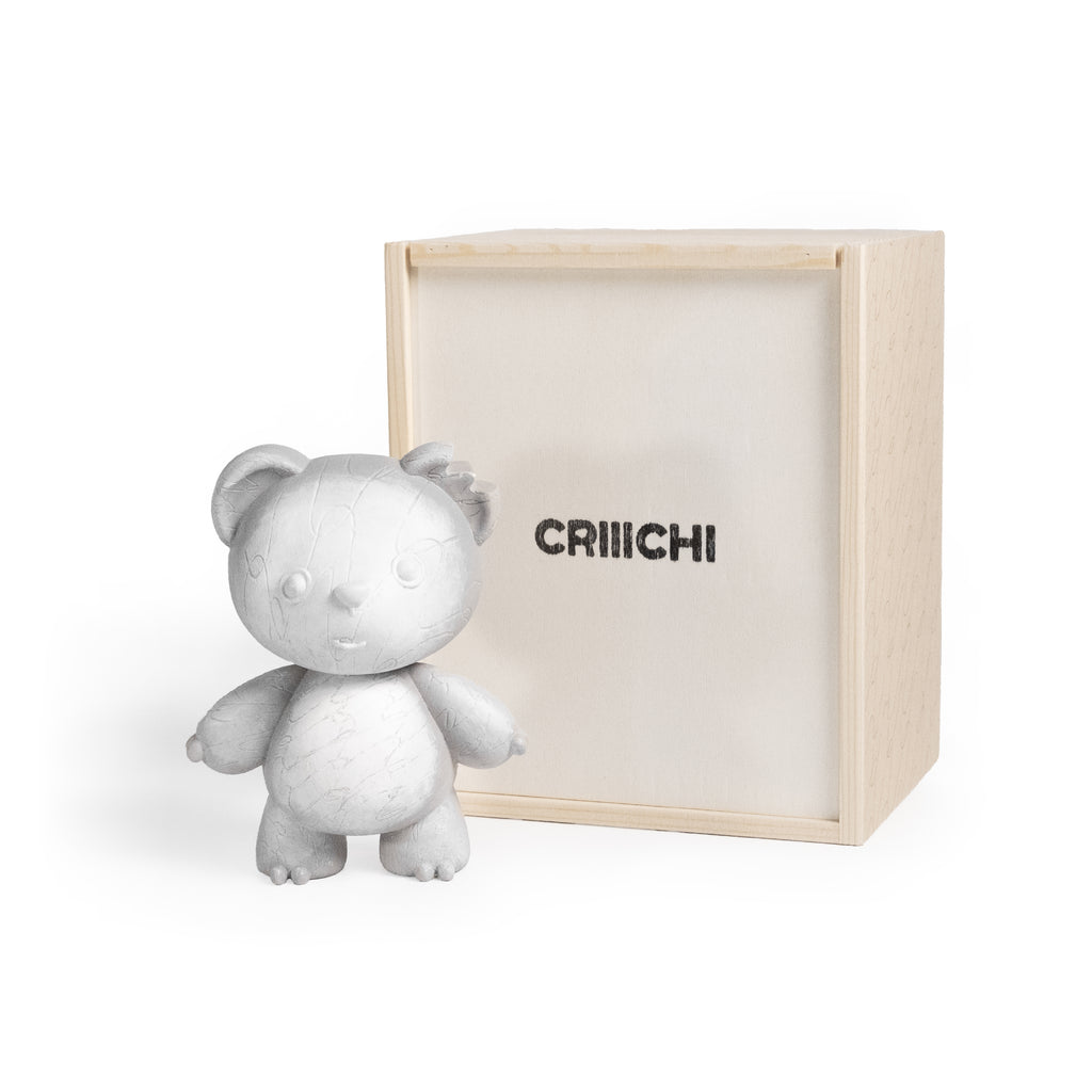 A light gray textured teddy bear figurine standing next to its natural wood presentation box, which features the brand name 'CRIIICHI' printed in black on the front.