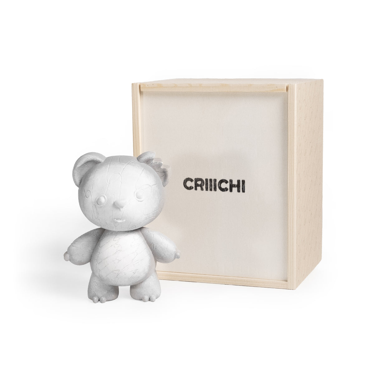 A light gray textured teddy bear figurine standing next to its natural wood presentation box, which features the brand name 'CRIIICHI' printed in black on the front.