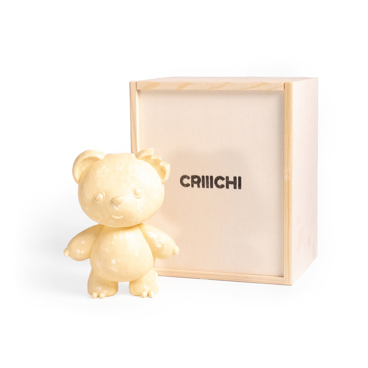 A pale cream-colored ceramic teddy bear figurine standing next to its light natural wood presentation box, which features the brand name 'CRIIICHI' printed in black.
