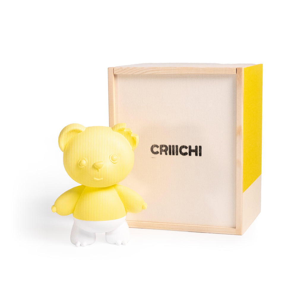 A yellow ribbed bear-shaped figurine with white pants stands in front of its light wood and yellow box packaging, which features the brand name 'CRIIICHI' printed on the front.