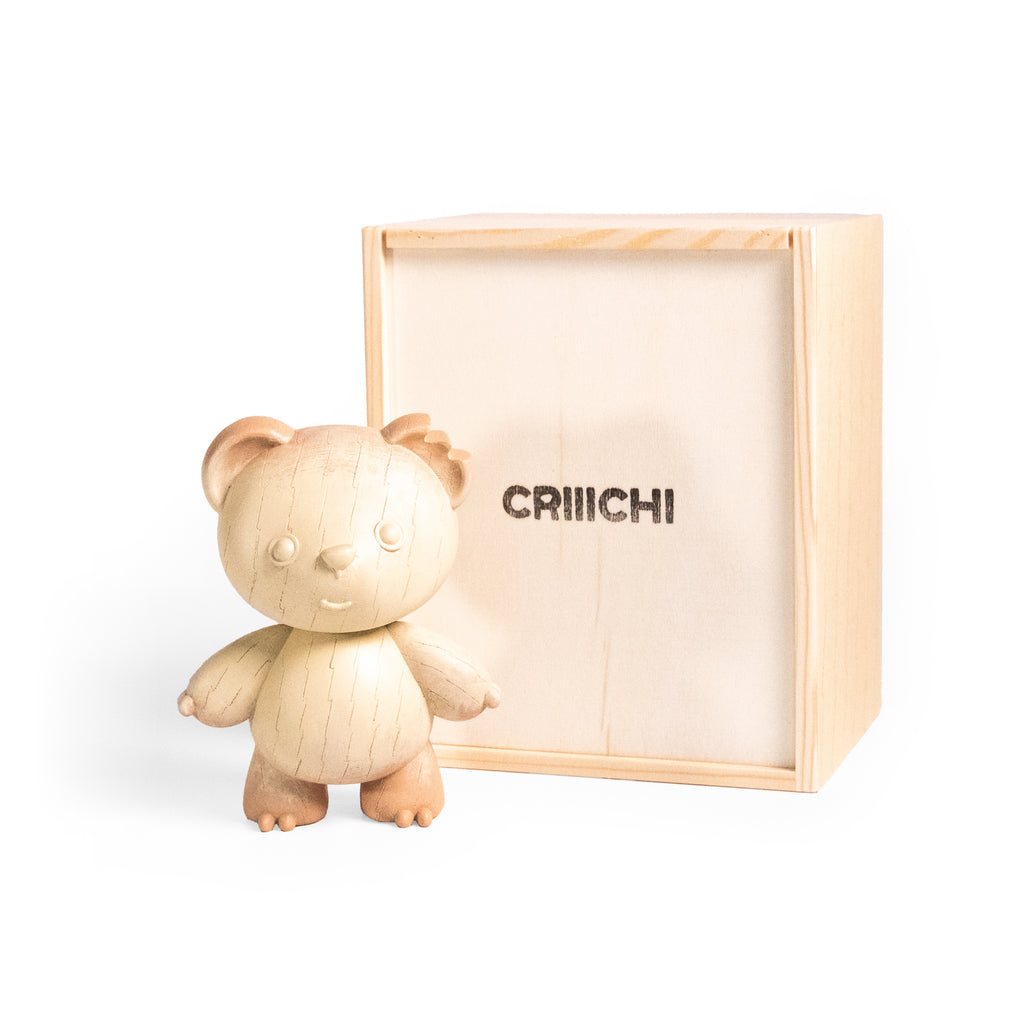 A small carved wooden teddy bear figurine standing in front of its light natural wood presentation box, which has the brand name 'CRIIICHI' printed on the front, set against a clean white background.
