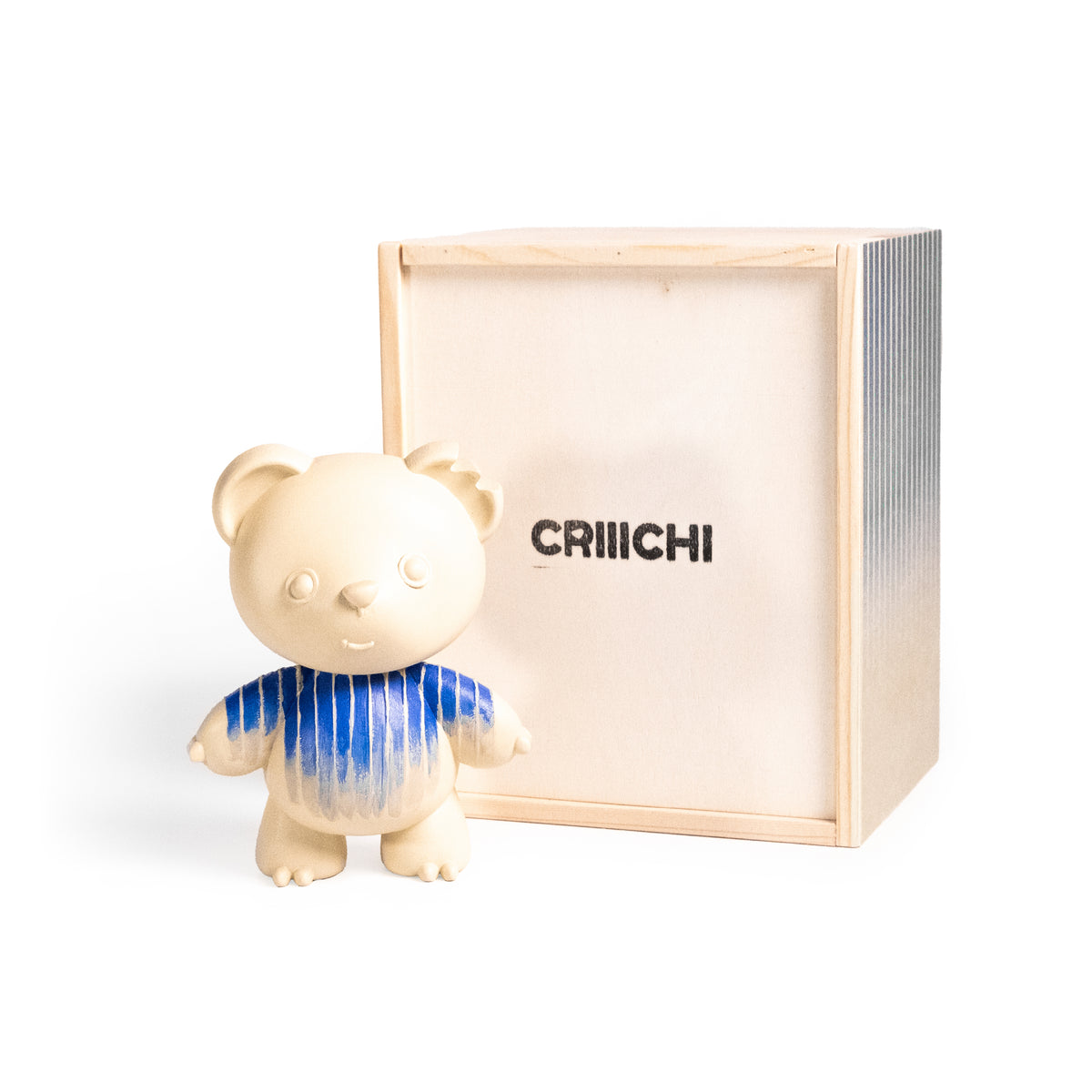 A cream-colored bear figurine wearing a blue striped shirt stands in front of its light wood packaging box, which features the brand name 'CRIIICHI' printed on the front.