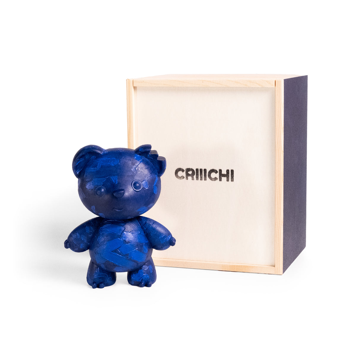 A small blue bear figurine with a textured, cracked surface stands in front of its light wood and dark navy presentation box, which has the brand name 'CRIIICHI' printed on the front, set against a plain white background.
