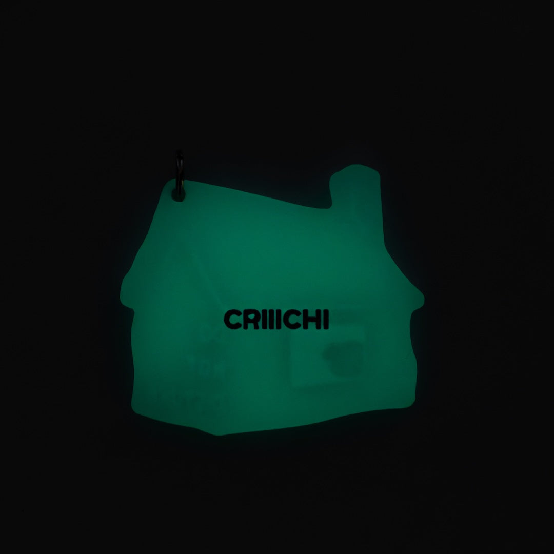 A glowing green house-shaped keychain with the brand name 'CRIIICHI' printed in black across the center, shown against a pitch-black background.