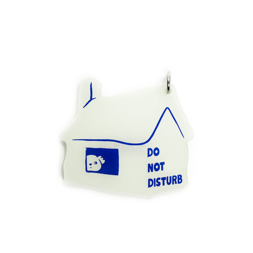 A small house-shaped pendant or charm in white with blue line art, a blue window graphic with a small face, and the words 'DO NOT DISTURB' printed in blue, shown on a plain white background.