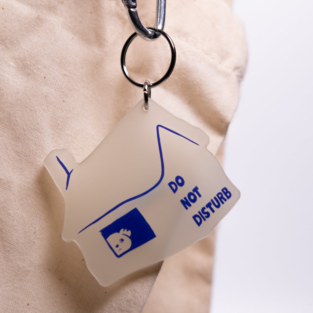 A translucent frosted acrylic keychain shaped like a house, featuring blue line art, a small blue square icon, and the text 'DO NOT DISTURB'. The keychain is attached to a black metal carabiner and is shown hanging against a beige textured fabric background.